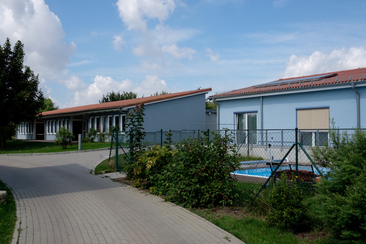 Die zwei einstöckigen Gebäude der Fördergruppe vor einem blauen Himmel. Im Vordergrund sieht man ein Schwimmbecken.