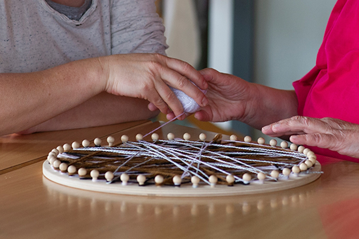 Zwei Personen arbeiten an einem runden Rahmen mit Stofffäden, der auf einem Tisch liegt.