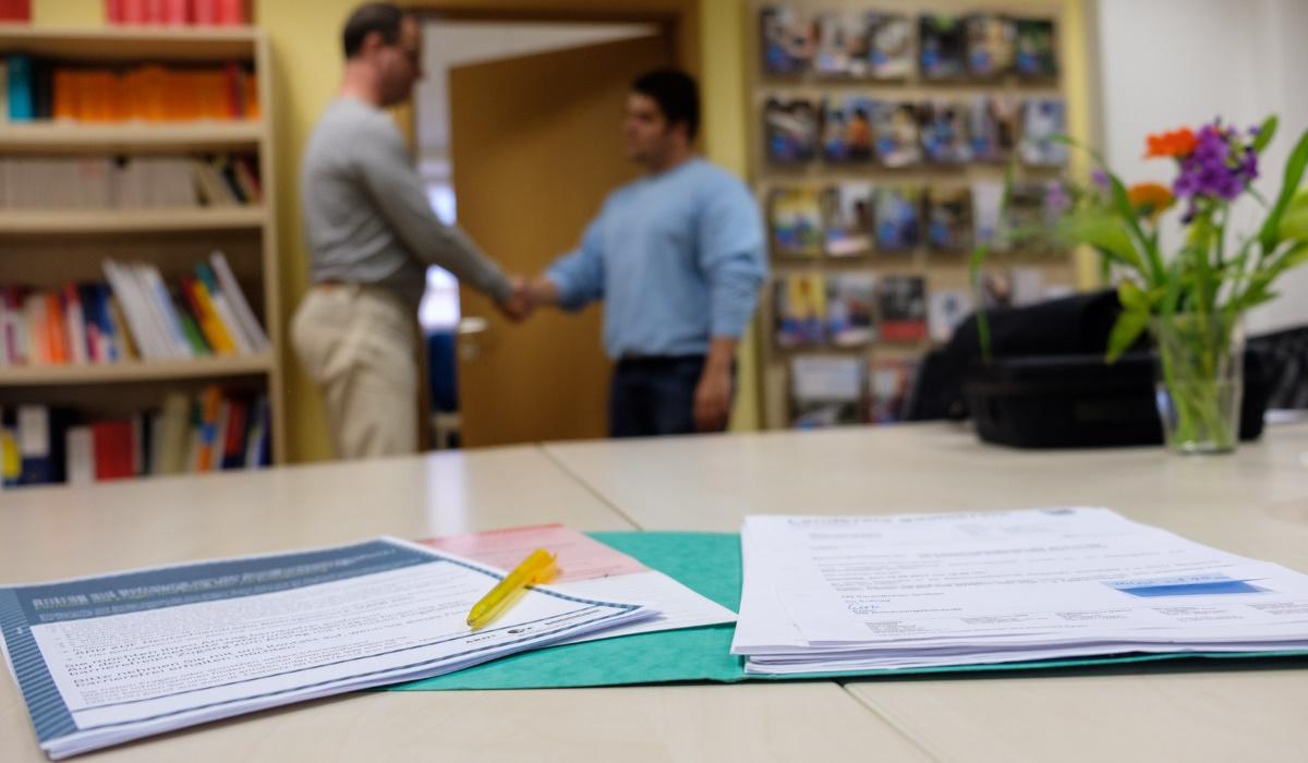 Schreibtisch im Vordergrund, zwei Menschen reichen sich im Hintergrund die Hand